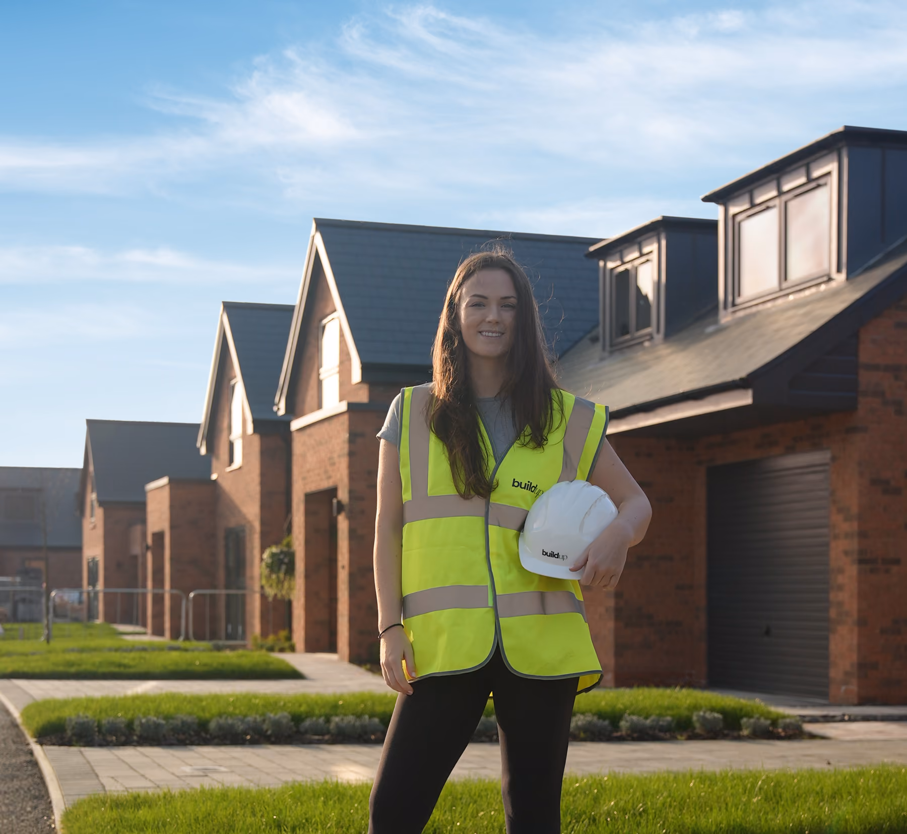 Rosey Cassidy in yellow safety vest holding white hardhat standing in front of modern brick houses on sunny day.