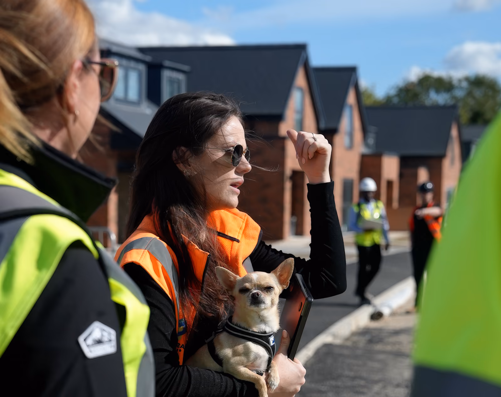 Rosey Cassidy in orange safety vest holding a small dog and gesturing amid construction workers and houses.